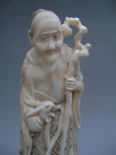 Ivory okimono depicting a tall Sage standing on a rock, with a long gnarled stick and holding a rabbit against him. Some lines of age, weak patina, signature under the base GYOKUSEN, origin Japan around 1910